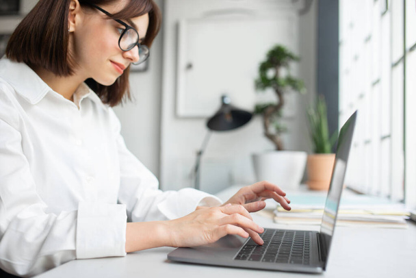 Young caucasian businesswoman in glasses working on computer, enjoying professional occupation in office interior, selective focus on hands on keyboard. Business, official lifestyle, ad and offer - Photo, Image