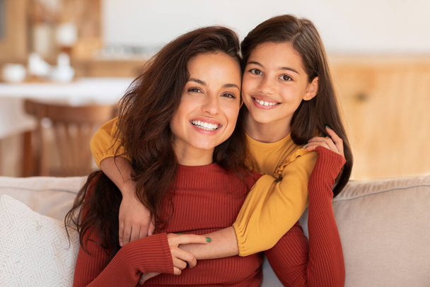 Mütter lieben. Porträt der glücklichen jungen Araberin und ihrer hübschen Tochter, die auf der Couch zu Hause posiert und in die Kamera lächelt. Liebevoll mutter bonding mit kind mädchen embracing drinnen - Foto, Bild