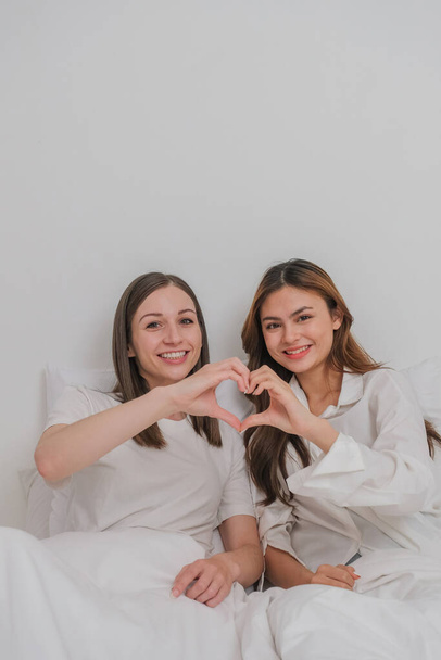 Lesbian couple together concept. Portrait of a smiling young lesbian couple other while sitting together and hand made heart symbol in the morning at bedroom. - Photo, Image