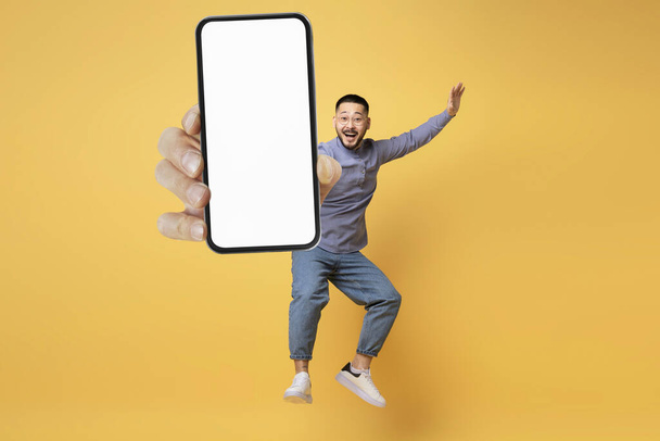 Tolles Angebot. Überraschter asiatischer Mann springt mit großem leeren Smartphone in der Hand über gelbem Hintergrund, aufgeregter Millennial Guy demonstriert leeres Handy mit weißem Bildschirm vor der Kamera, Mockup - Foto, Bild