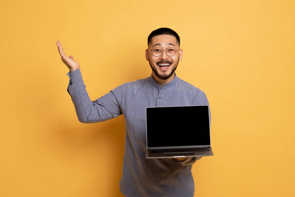 Великий Озил. Возбужденный азиат держит ноутбук компьютер с чистым экраном и восклицает с радостью, удивленный парень тысячелетия показывает копировать пространство для онлайн-предложения, стоя на желтом фоне, макет - Фото, изображение