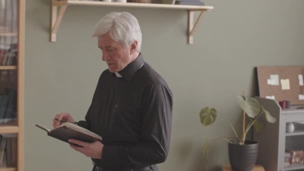 Portrait moyen d'un prêtre caucasien senior en chemise noire et col clérical prenant des notes dans un carnet puis souriant à la caméra, travaillant depuis son bureau à domicile - Séquence, vidéo