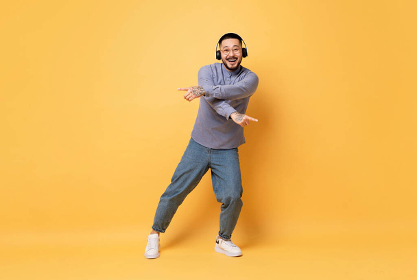 Carefree Young Asian Guy Wearing Wireless Headphones Dancing On Yellow Background, Happy Millennial Man In Eyeglasses Listening Music, Enjoying Favorite Songs And Having Fun In Studio, Fuul Length - Foto, Imagen