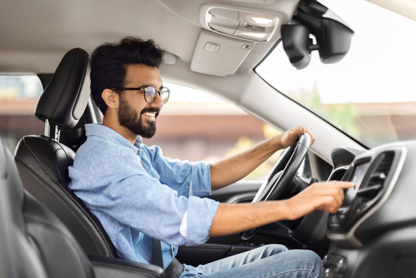 Soittolista. Side näkymä onnellinen nuori intialainen mies ajaa autoa ja kuunnella musiikkia radio auto audiojärjestelmä, tarkkailun auto vaihtoehtoja koeajon aikana, painike kojelautaan - Valokuva, kuva