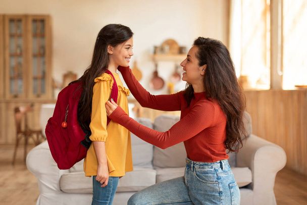 Πίσω στο σχολείο. Ευτυχισμένη μαμά της Μέσης Ανατολής προετοιμασία προεφηβική κόρη μαθήτρια με σακίδιο για την πρώτη σχολική μέρα στο σπίτι, κοιτάζοντας το παιδί και ενθαρρύνοντάς την να απολαύσετε τα μαθήματα το πρωί - Φωτογραφία, εικόνα