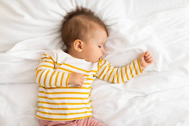 Adorable little baby girl sleeping on white bedding, feeling safe, watching sweet dreams, growing, gaining strength, top view, copy space. Peaceful child napping during daytime sleep - Photo, Image