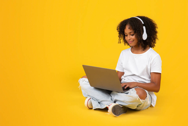 Vrolijke puber Afro-Amerikaanse meisje in wit t-shirt, draadloze hoofdtelefoon met computer, kijken online les geïsoleerd op oranje achtergrond, studio. Huiswerk met apparatuur, modern onderwijs - Foto, afbeelding