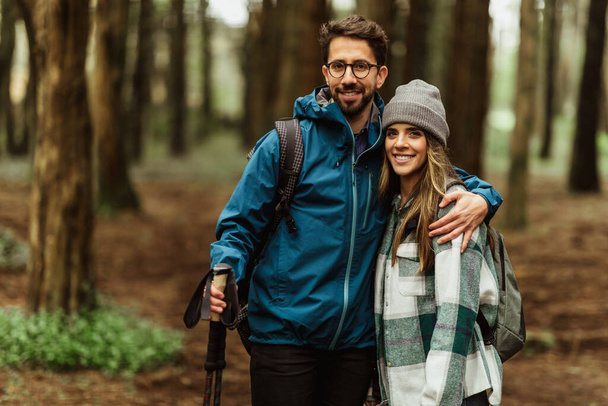 Positive young caucasian couple in jackets with trekking sticks walk in forest together, enjoy trip, active lifestyle, outdoor. Travel hiking, adventure and tourism, relationships - Photo, Image