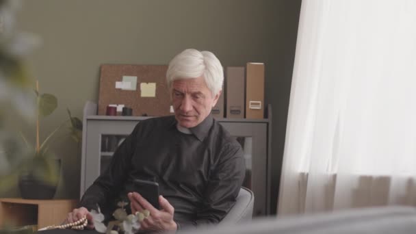 Senior ősz hajú pap visel fekete ing és fehér papi gallér, hogy az online video beszélgetés egyházközség okostelefon, dolgozik otthonról - Felvétel, videó
