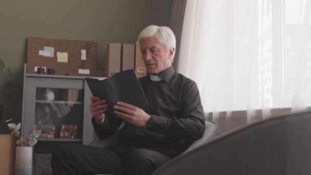 Sacerdote senior en camisa negra y cuello clerical blanco leyendo la Biblia en voz alta en casa - Metraje, vídeo