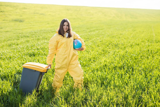 Sarı koruyucu takım elbiseli bir kadın yeşil bir tarlanın ortasında elinde bir küre tutarken diğer eliyle bir çöp tenekesi taşıyor.. - Fotoğraf, Görsel