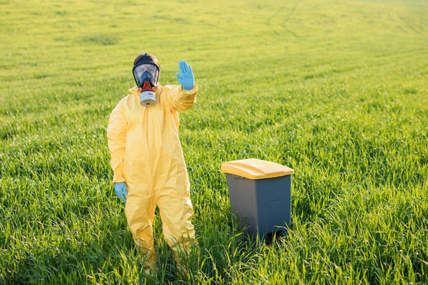 Eine Frau in einem gelben Schutzanzug steht in einer schützenden Gasmaske inmitten eines grünen Feldes und zeigt eine Geste, um zum Anhalten und zur Rettung des Planeten aufzurufen. Eine Frau steht neben der Mülltonne und ruft dazu auf, den Planeten zu retten - Foto, Bild