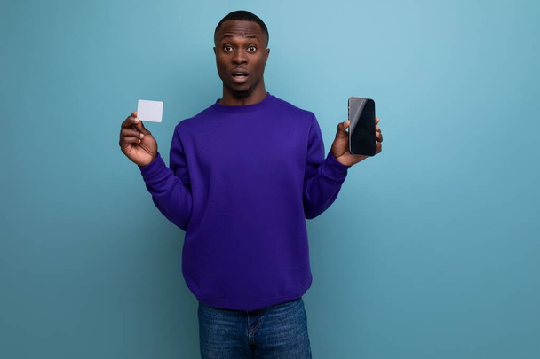 guapo joven africano en azul sudadera con capucha utiliza teléfono y tarjeta bancaria. - Foto, imagen