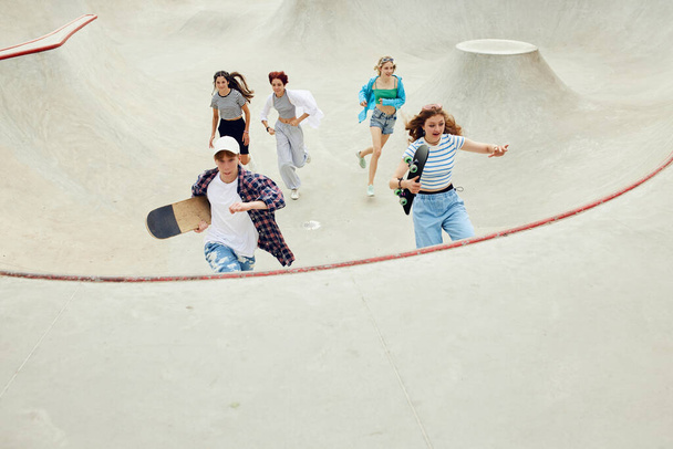 Grupo de adolescentes en ropa casual, niños y niñas corriendo con patín en rampa de skate. Actividad y diversión. Concepto de cultura juvenil, deporte, dinámica, extrema, afición, acción y movimientos, amistad - Foto, imagen