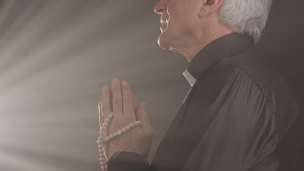 Hitaasti vanhempi pappi rukoilee kädet puhuen Jumalalle, katsoen kirkasta valoa pimeässä kirkossa - Materiaali, video