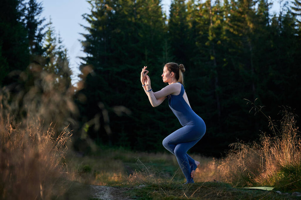 Jeune femme pratiquant des poses de yoga dans la forêt de conifères. Femme d'âge moyen faisant la pose d'aigle. Femme en survêtement bleu foncé s'exerçant le matin. - Photo, image