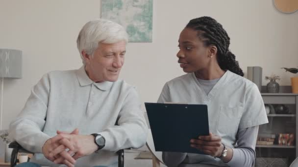 Portrait of cheerful Caucasian old man in wheelchair and young African American female nurse with patient medical card on clipboard in hands smiling at camera sitting at mans home indoors - Footage, Video