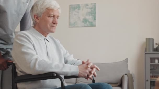 Pielęgniarka zajmująca się białym seniorem siedzącym w domu na wózku inwalidzkim, otwierającym mu laptopa i przynoszącym szklankę wody - Materiał filmowy, wideo