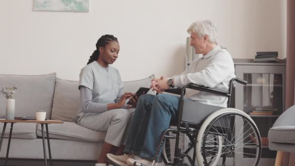 Genç Afrikalı Amerikalı kadın sosyal hizmetler görevlisi evinde tekerlekli sandalyede oturan beyaz yaşlı adama dijital tableti nasıl kullanacağını anlatıyor. - Video, Çekim