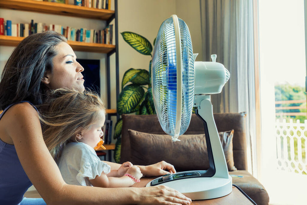 Mother and daughter make cooling off with a fan in the living room a fun dance game, forgetting the heat, seeking relief, and having fun - Photo, Image