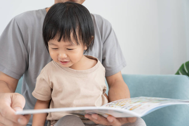 Mutlu Asyalı baba rahatla ve evde bebek zamanlarıyla kitap oku. Ebeveyn kızıyla kanepede oturup masal okuyor. öğrenme, çocuk bakımı, gülme, eğitim, hikaye anlatma, uygulama. - Fotoğraf, Görsel