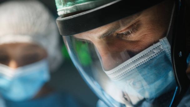 Cirujano con visera y máscara quirúrgica opera una enfermedad contagiosa. - Foto, Imagen