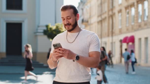 Uśmiechnięty brodaty młody człowiek za pomocą smartfona wpisując wiadomości tekstowe przeglądając internet, kończąc pracę, patrząc na kamery na zewnątrz. Facet turysta spacerujący ulicą miejskiego słońca. Styl życia miasta - Materiał filmowy, wideo