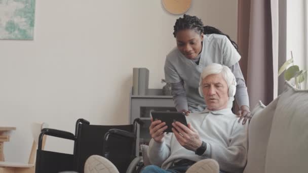 Hombre mayor caucásico en auriculares inalámbricos sentado en el sofá en su apartamento aprendiendo a usar la tableta digital, mientras que la trabajadora social afroamericana lo ayuda - Imágenes, Vídeo