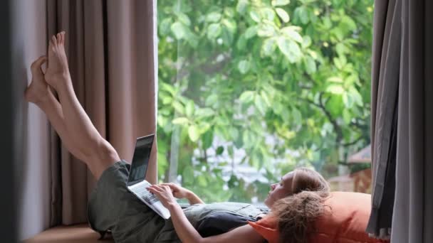 Fiatal lány fekszik az ablakpárkányon közel nagy ablak és csevegés barátaival keresztül video chat segítségével egy számítógép laptop.  - Felvétel, videó