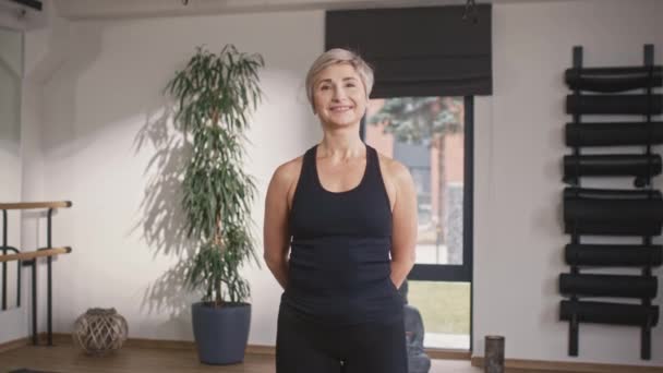 Positive Caucasian woman wearing black sport clothes standing in front of camera and happilly smiling. Feeling wellness after meditation in fitness room. Healthy lifestyle concept. - Footage, Video