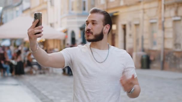 Parrakas nuori mies bloggaaja ottaa selfie älypuhelimeen, kommunikoida videopuhelu verkossa tilaajien kanssa, tallennus tarinoita sosiaalisen median vlog ulkona. Guy kävely kaupunkien auringonpaiste kaupunki katu - Materiaali, video