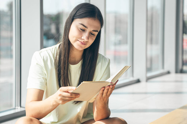 Houkutteleva nuori nainen lukee kirjaa istuessaan ikkunan ääressä kesällä aurinkoisena päivänä, kopioi tilaa. - Valokuva, kuva