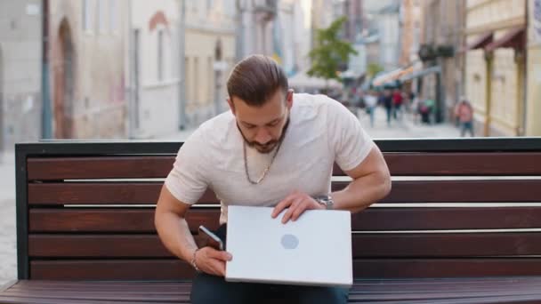 Portret van een jonge gelukkige man opent een laptop zittend op een bankje in de stad in de buitenlucht. Midden-Oosten kerel werkt op notebook, stuurt berichten, maakt online aankopen, kijkt films. Stadslevensstijl - Video