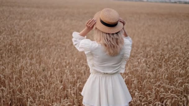 Retrato de uma jovem loira usando um chapéu de palha e se voltando para a câmera com um sorriso encantador enquanto caminha em um campo de trigo dourado em um dia ensolarado - Filmagem, Vídeo