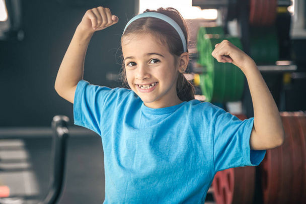 Charmantes kleines Mädchen zeigt ihren Bizeps und lächelt, während sie im Fitnessstudio trainiert. - Foto, Bild
