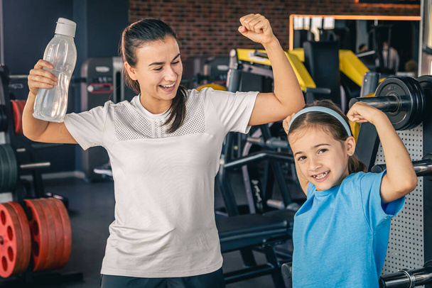 Piękna silna kobieta i urocza córeczka pokazują swoje bicepsy i uśmiechają się podczas treningu na siłowni. - Zdjęcie, obraz