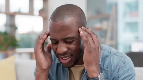 Stress, hoofdpijn en zwarte man op de bank met tempelmassage voor hersenmist in zijn huis. Angst, migraine en Afrikaanse man gefrustreerd door duizeligheid, zorgen of geestelijke gezondheid, zorgen of crisis in de woonkamer. - Video