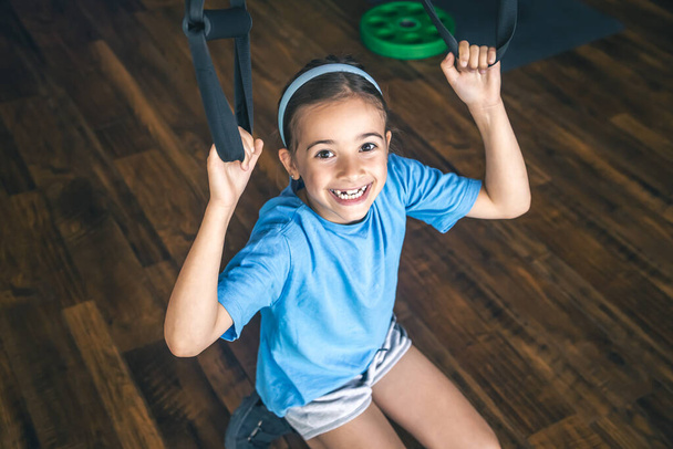 Little girl exercising indoors using trx fitness straps for working with his own weight, doing squats or training upper body. - Photo, Image