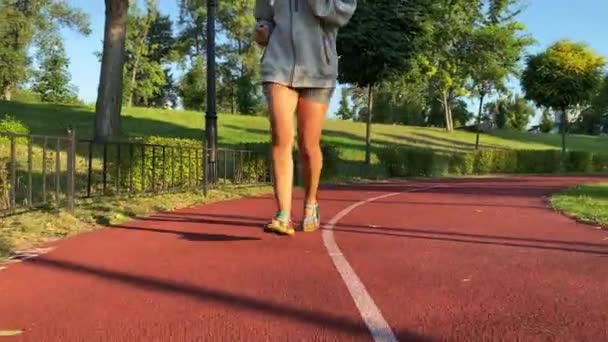 Wielorasowa młoda kobieta idzie na poranny jogging po szlaku w parku publicznym. Biegaczka biegnie skąpana w złocistym słońcu na tle przejrzystego błękitnego nieba i miejskiego krajobrazu. Sprawność miejska, witalność - Materiał filmowy, wideo