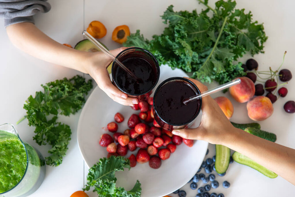 Mutter und Kind halten Gläser mit Beeren-Smoothies in der Hand vor dem Hintergrund eines Küchentisches mit Gemüse und Obst. - Foto, Bild