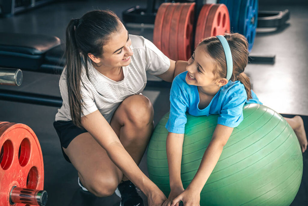 Маленькая спортивная девочка и молодая красивая мама делают растяжку с фитнес-мяч в тренажерном зале. - Фото, изображение
