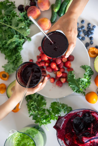 Mom and child hold glasses with berry smoothies in their hands against the background of a kitchen table with vegetables and fruits. - Photo, Image