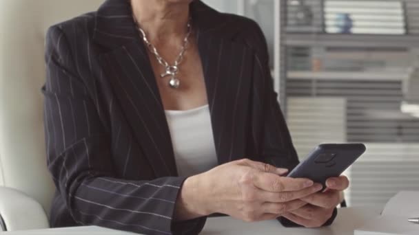 Приглушений знімок серйозної зрілої азіатської бізнес-леді в смугастій куртці та окулярах, використовуючи свій смартфон на робочому місці - Кадри, відео