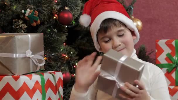 Caucasian little boy shaking the christmas gift box. Male kid trying to know what inside the gift box near the christmas tree. Little boy examining silver colored gift box - Footage, Video