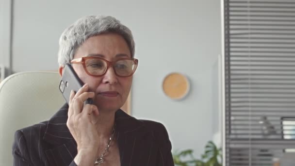 Zelfverzekerde middelbare leeftijd Aziatische zakenvrouw praten op mobiele telefoon terwijl zitten aan het bureau in haar moderne kantoor - Video