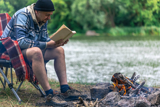 Close-up van het kampvuur met een man die een boek leest op de achtergrond in de natuur, outdoor recreatie concept, kopieer ruimte. - Foto, afbeelding