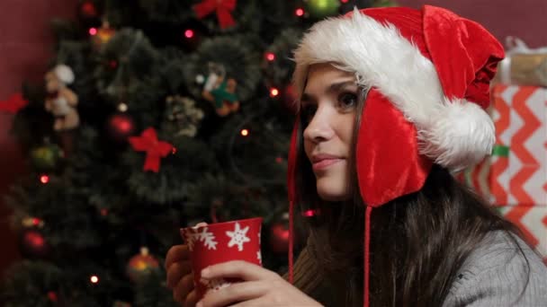 Jovem caucasiana de chapéu de Pai Natal a beber uma bebida quente. Mulher atraente segurando copo vermelho contra fundo da árvore de Natal. Mulher morena bonita olhando para copyspace perto do Natal
 - Filmagem, Vídeo