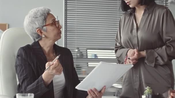 Mulher de negócios asiática madura em jaqueta listrada e óculos sentados no chefe da mesa de escritório durante a realização de reunião de equipe para discutir ideias do projeto e resultados da empresa - Filmagem, Vídeo