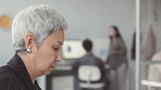 Side view van zelfverzekerde middelbare leeftijd Aziatische vrouw het maken van telefoongesprek tijdens het werken in hedendaags kantoor met haar collega 's chatten door bureau op de achtergrond - Video