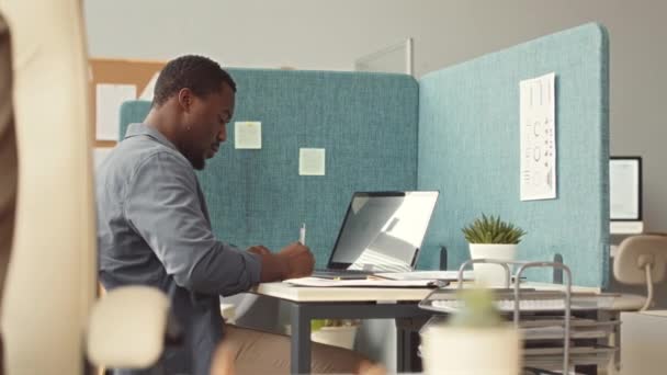 Сосредоточенный молодой афроамериканец пишет заметки на наклейках во время работы над ноутбуком в современном офисе - Кадры, видео